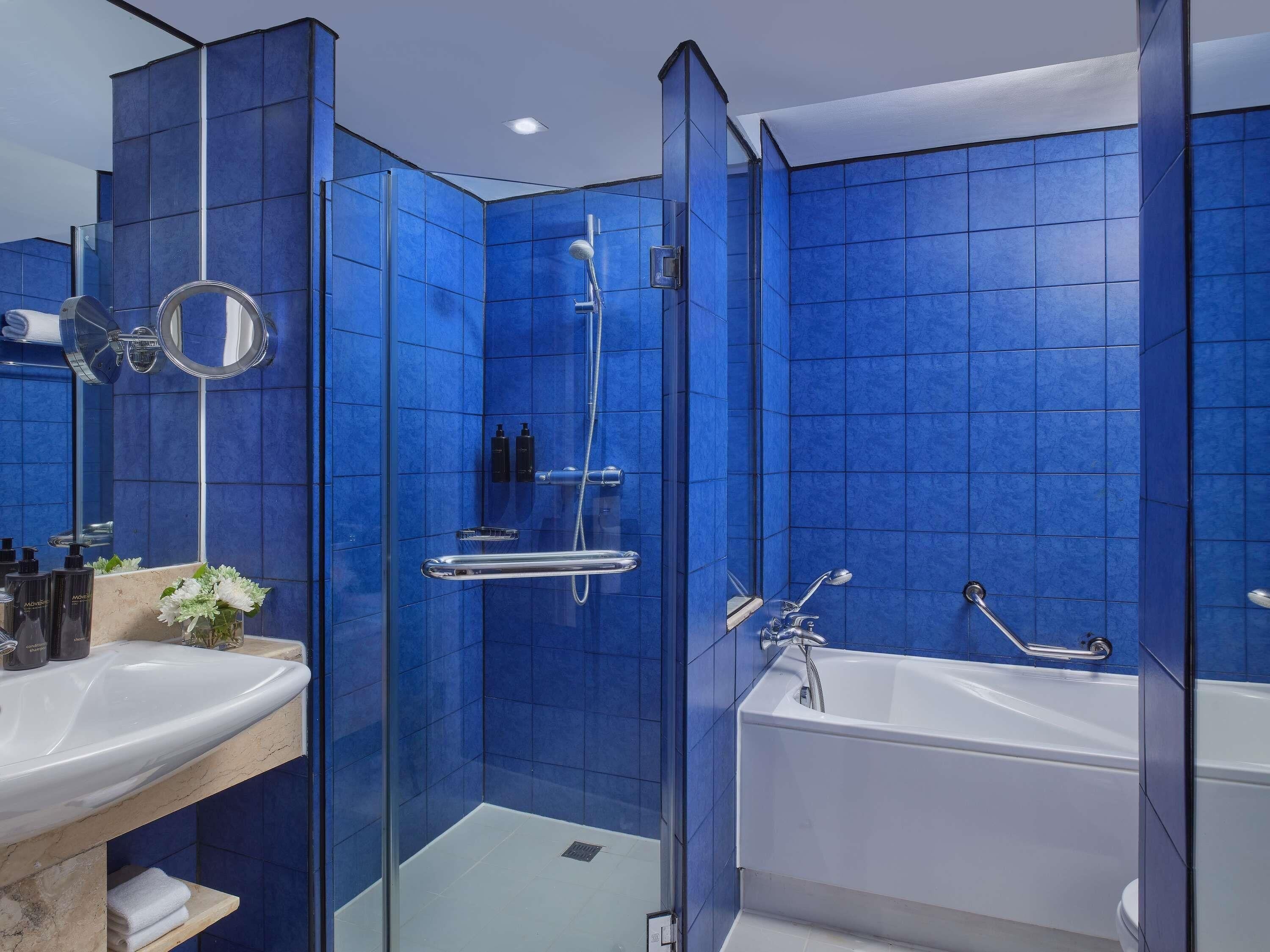A modern bathroom with blue tiles, a glass shower enclosure, a white bathtub, and a vanity with a mirror and toiletries.