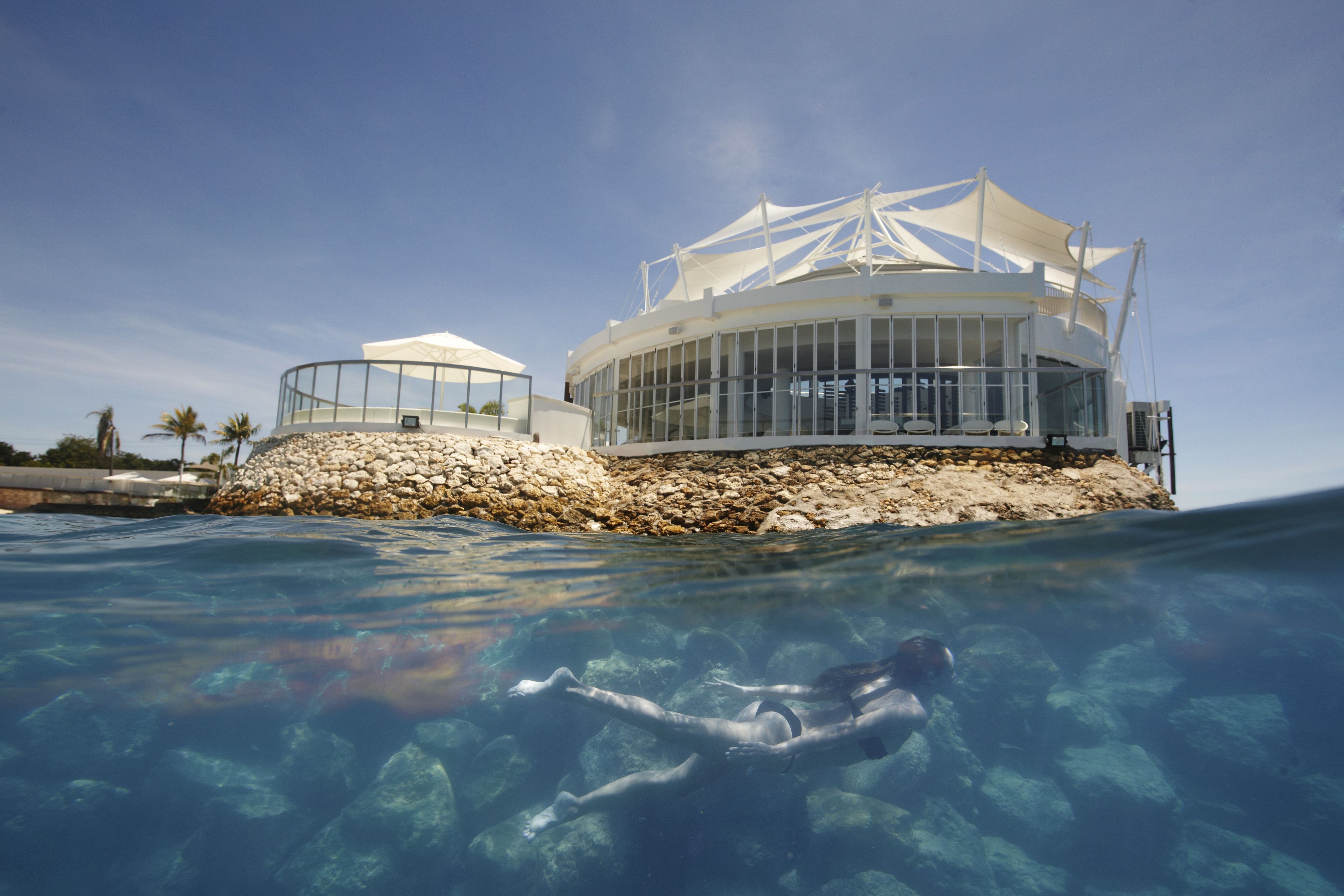 Edifici modern d'estructura circular i façana transparent, a prop d'una piscina cristal·lina amb roques visibles i un banyista.