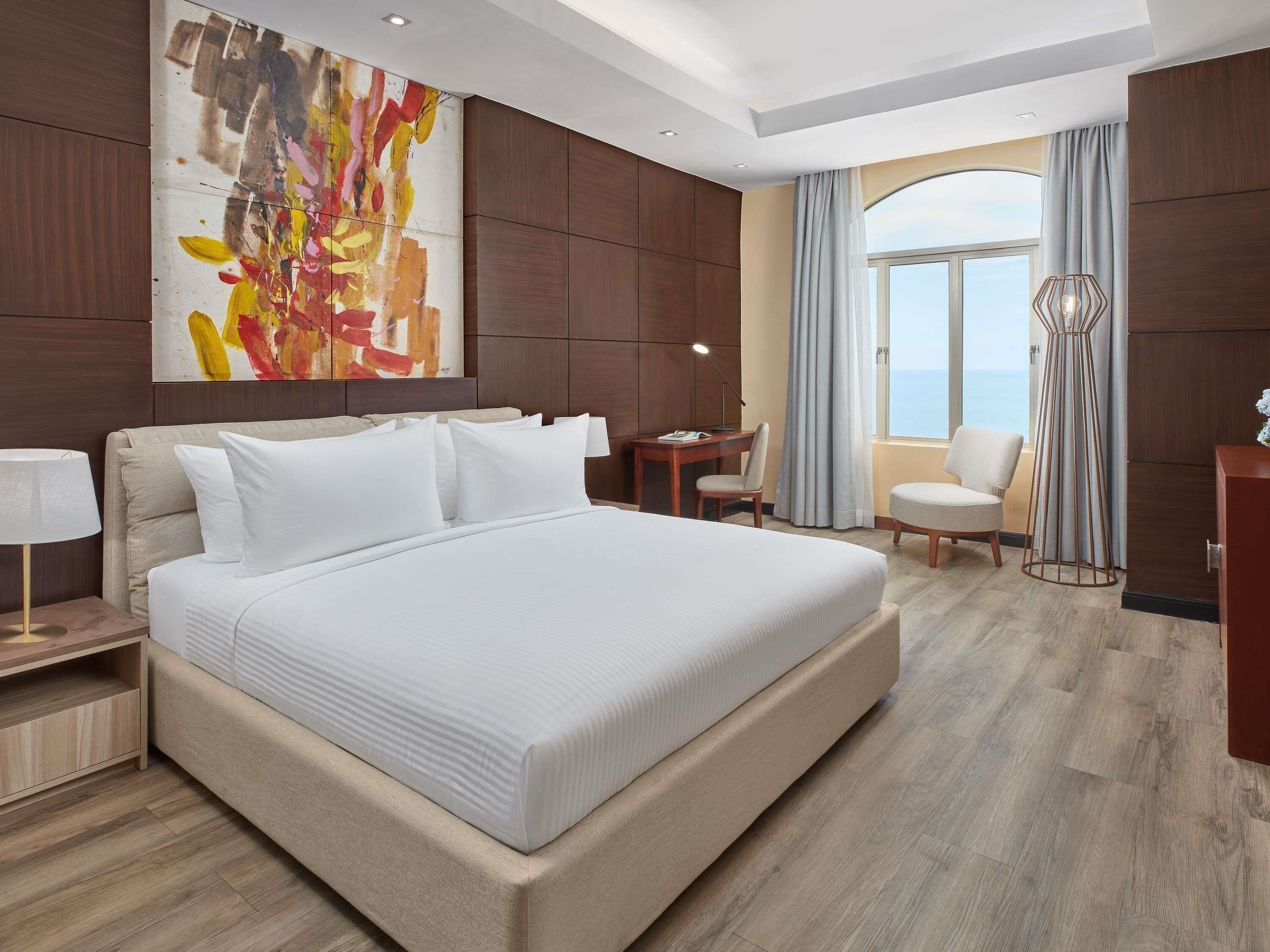 A modern hotel room with a large bed, a desk, a chair, and a view of the sea.