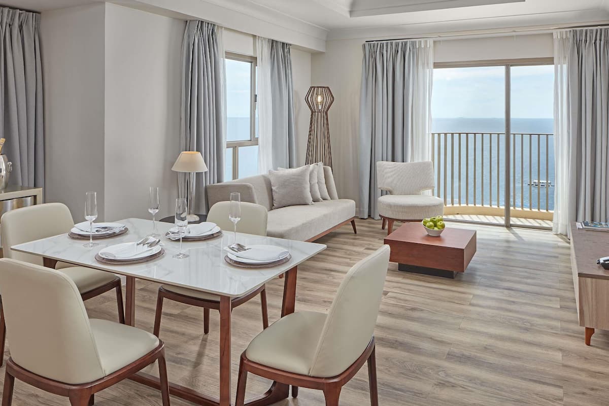 A modern hotel room with a dining area, a sofa, a chair, and a balcony with a view of the sea.