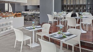 A modern restaurant with white wicker furniture, a buffet area, and a view of the kitchen. - Mövenpick Hotel Mactan Island Cebu (Lapu-Lapu)