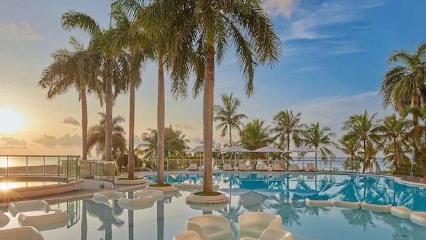 Un espace piscine avec des chaises longues, des palmiers et une vue sur l’océan au coucher du soleil.