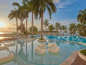 Un espace piscine avec des chaises longues, des palmiers et une vue sur l’océan au coucher du soleil.