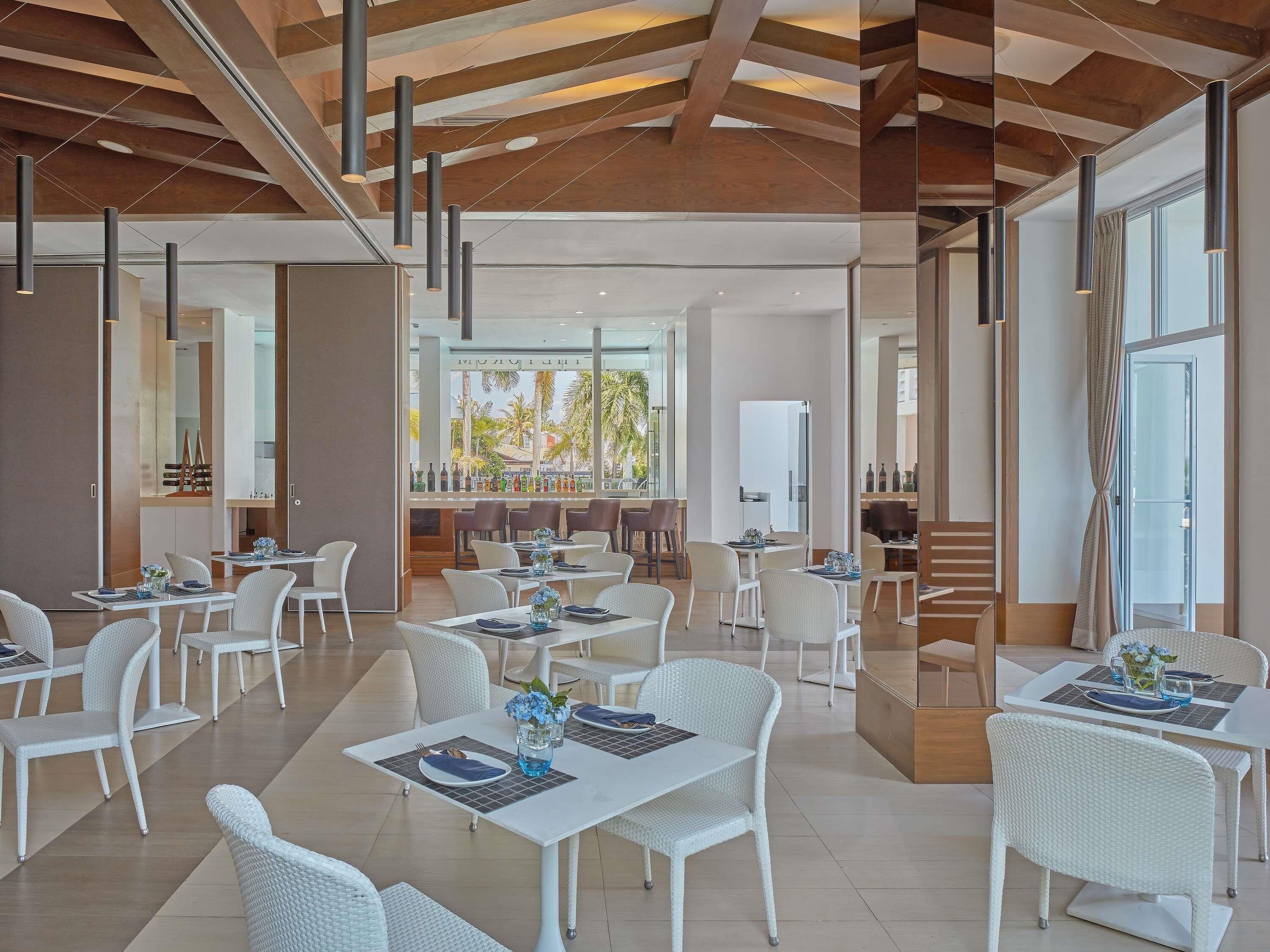 A modern dining area with white wicker furniture, pendant lighting, and a view of palm trees outside.