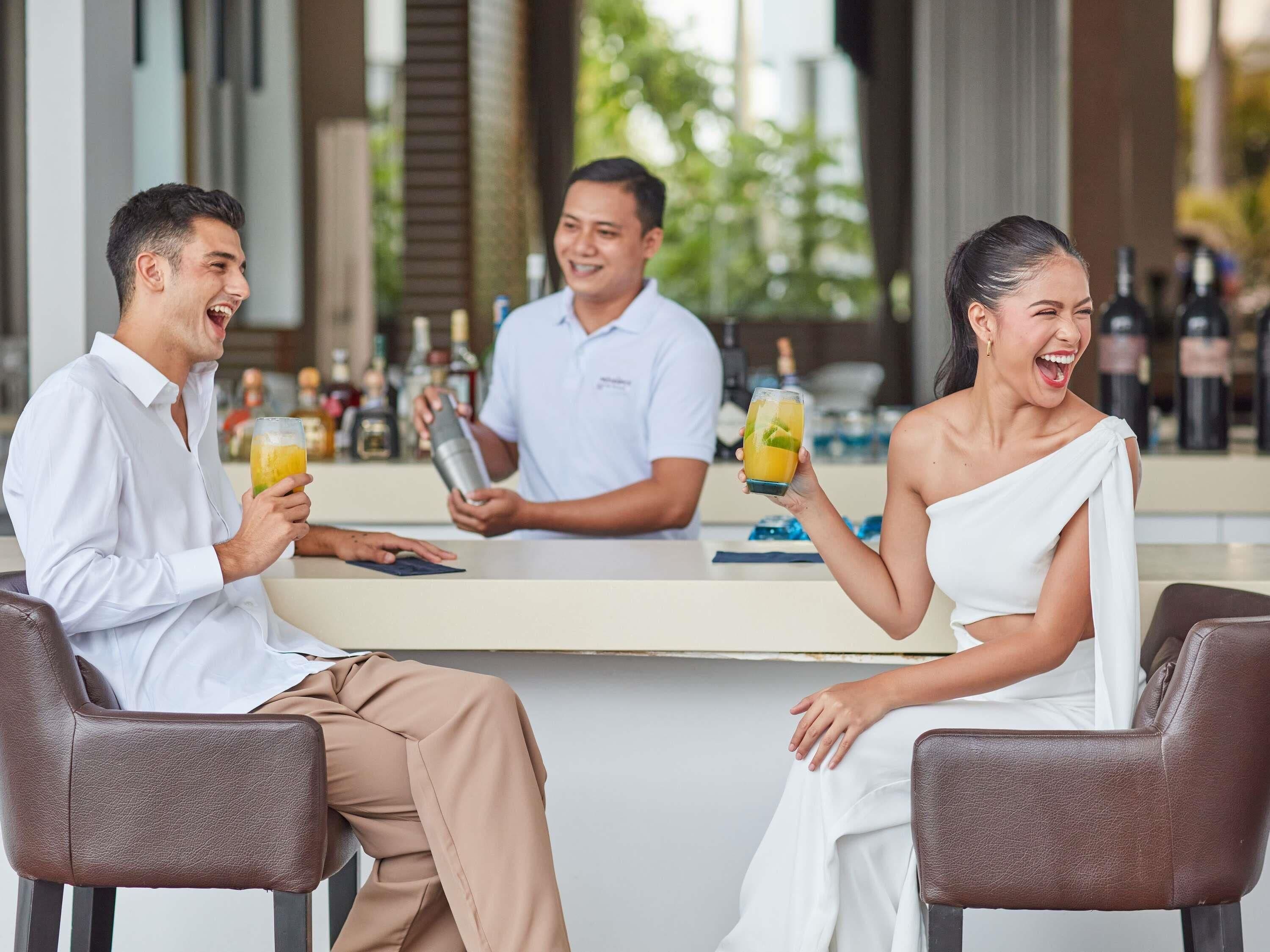 A bar area with three people seated at a counter, a bartender behind them, and various bottles of alcohol in the background.