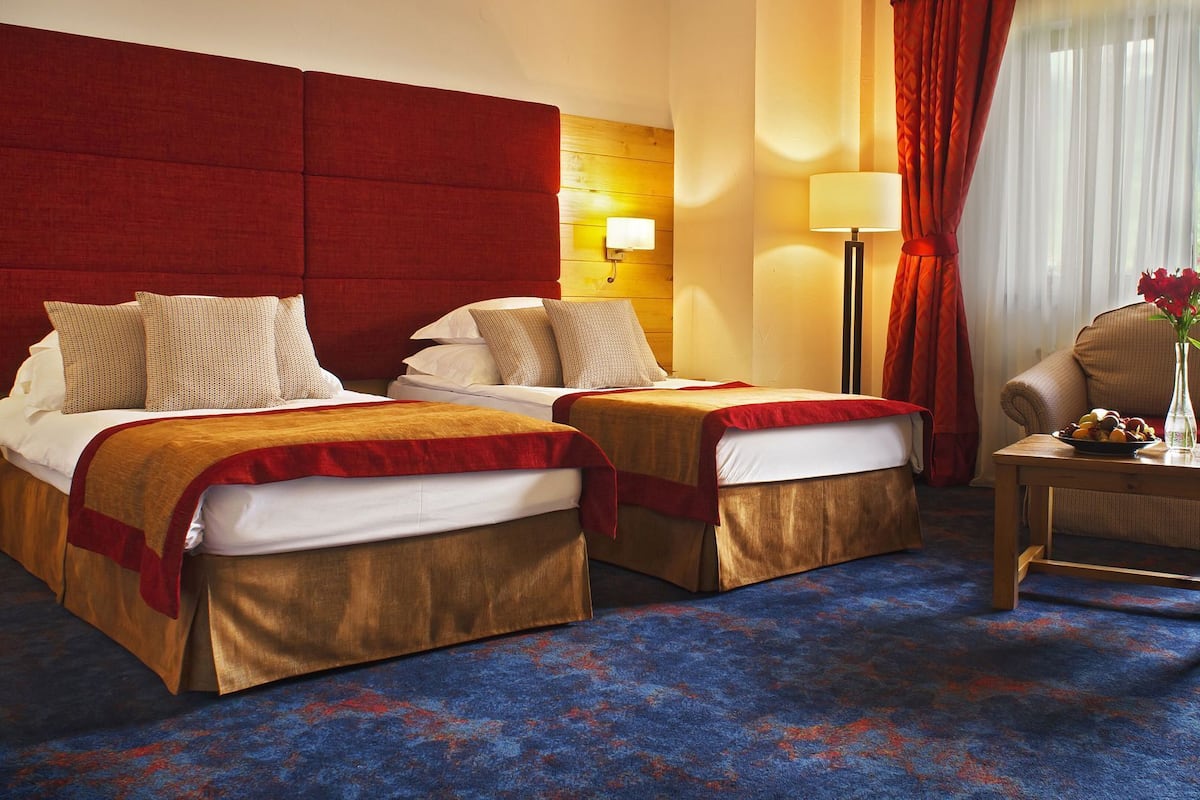 A hotel room with two beds, a wooden headboard, a nightstand, a lamp, a vase with flowers, and a view of the outdoors.