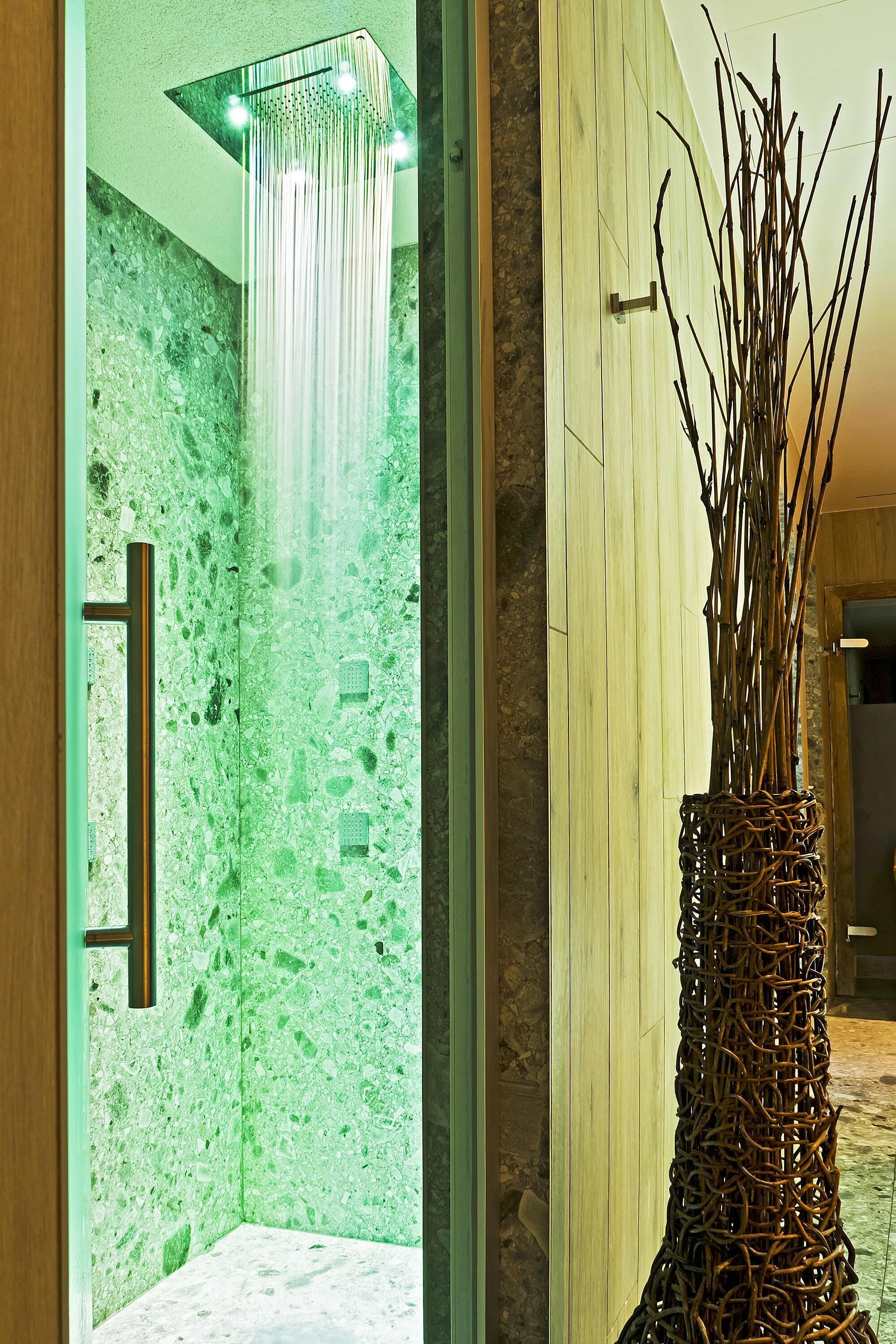 Un baño moderno con pared de mármol verde, ducha con puerta de vidrio y un jarrón decorativo con ramas.