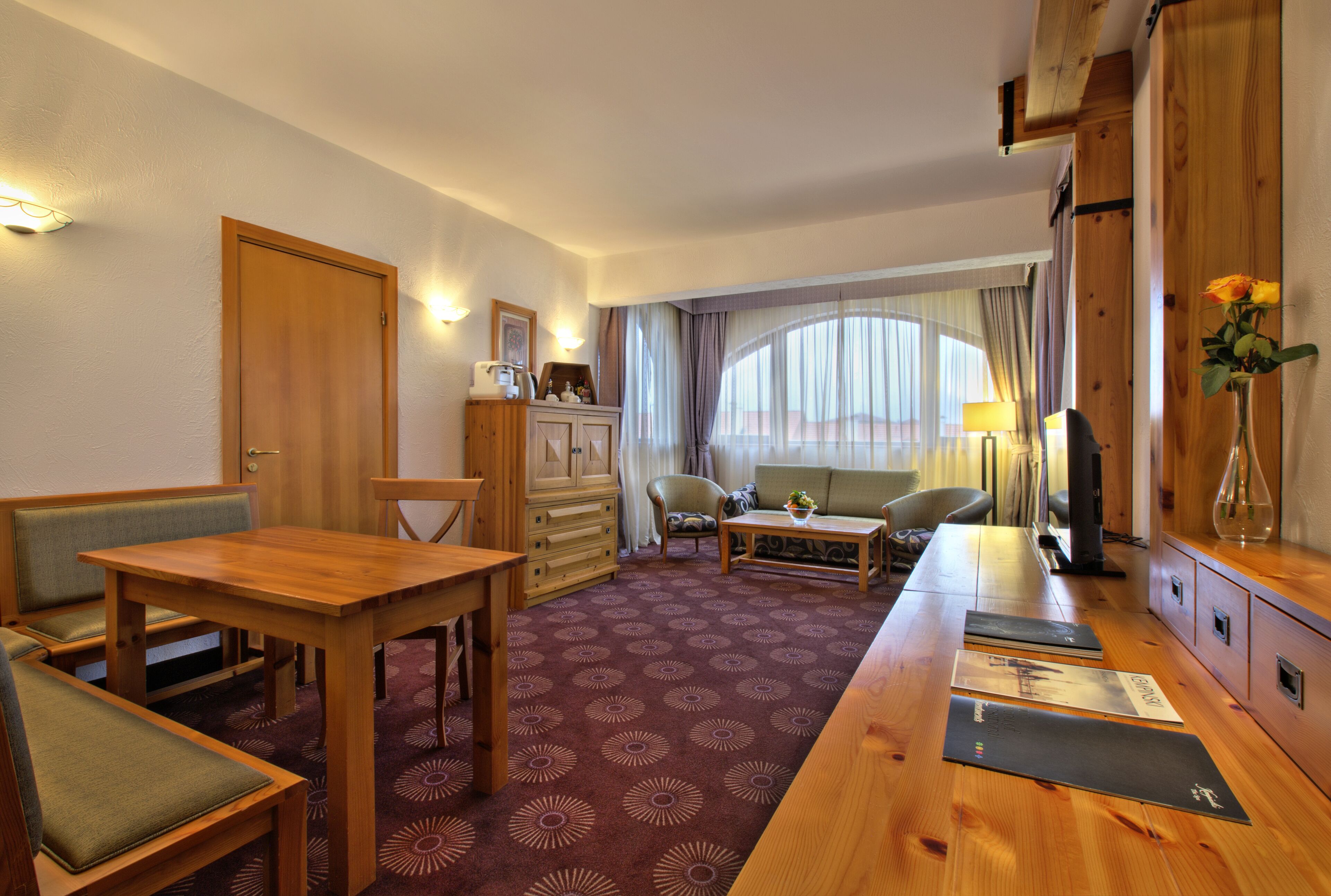 A hotel room with a wooden dining table, a sofa, a small armchair, a TV, and a desk with a lamp.