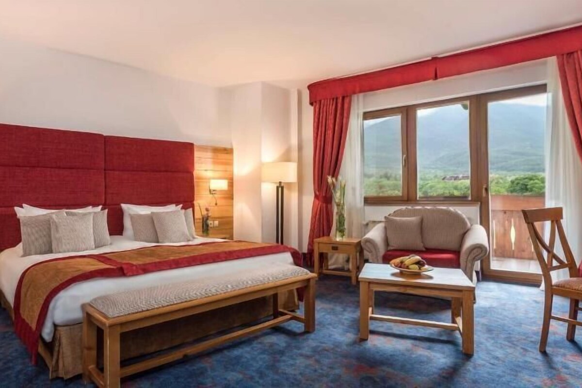 A hotel room with a large bed, a wooden nightstand, a bedside table, a chair, a small table with a bowl, and a window with curtains.