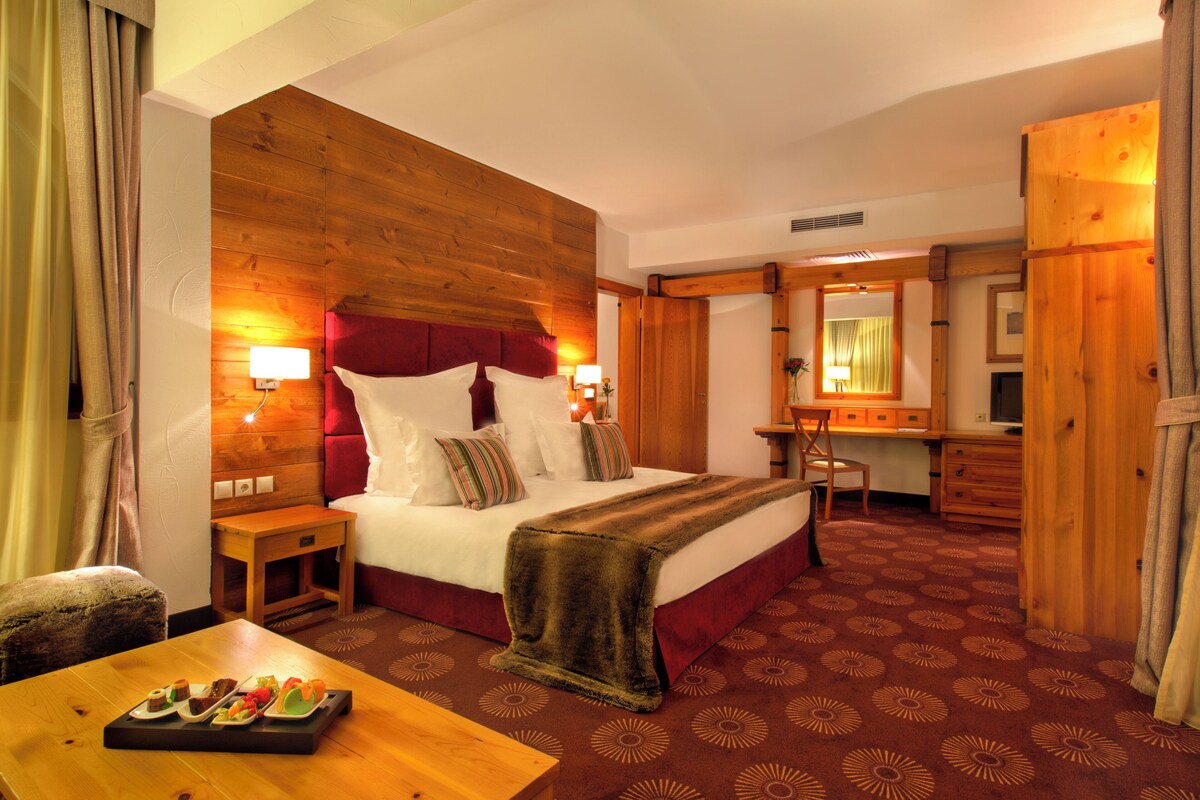 A hotel room with a wooden headboard, a bed with white bedding, a desk with a chair, a small table with pastries, and a patterned carpet.