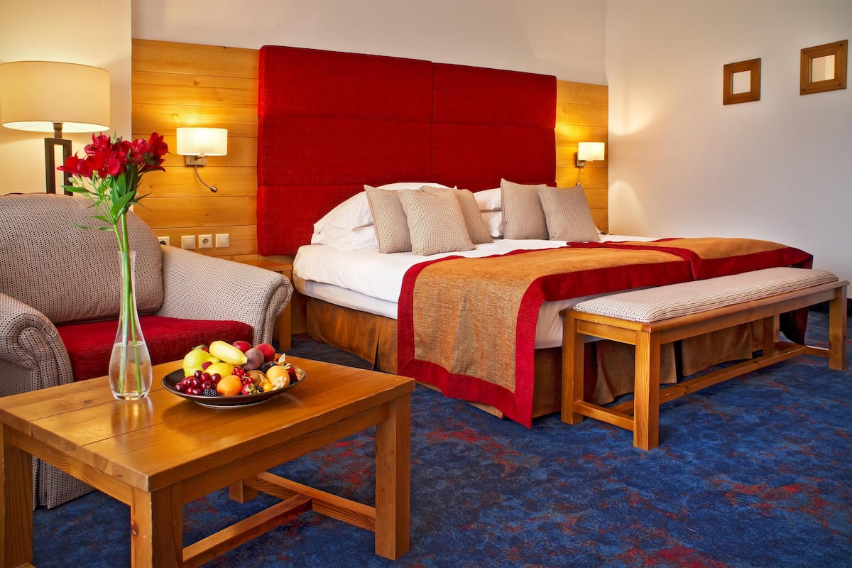 A hotel room with a large bed, a wooden bench, a dining table with fruit, and a vase of flowers.