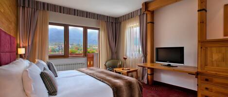 A hotel room with a large bed, a television, a desk, and a view of the mountains.