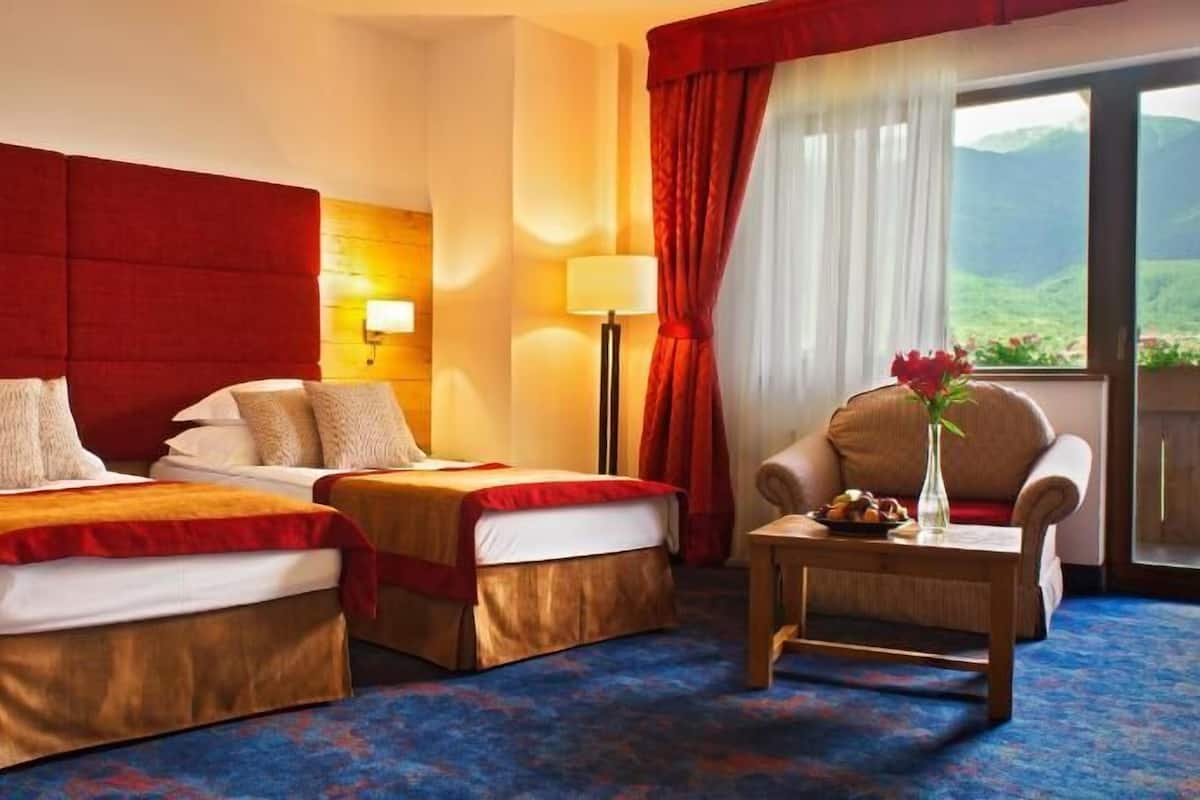 A hotel room with two beds, a chair, a small table, a lamp, and a view of the outdoors.