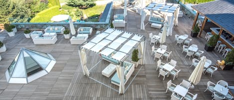 Un área de terraza en la azotea con varios conjuntos de mobiliario, incluyendo mesas y sillas, y un espacio lounge con paredes de vidrio.