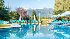 A pool with lounge chairs and umbrellas, surrounded by trees and mountains in the background. - Kempinski Hotel Grand Arena (Bansko)