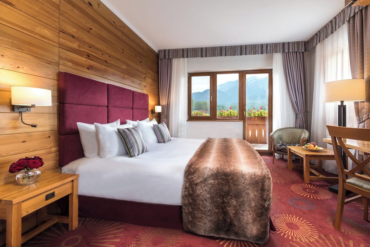 A hotel room with a wooden headboard, a large bed, a wooden nightstand with a vase of flowers, a wooden bench, a table with fruit, and a view of mountains through the window.