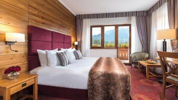 Habitación de hotel con cabecera de madera, una cama grande, una mesita de noche con un jarrón de flores, un banco de madera y una mesa con fruta, con vista a las montañas a través de la ventana.
