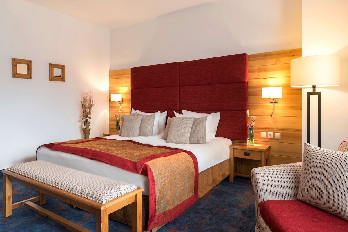 A hotel room with a large bed, bedside tables, a bench, a chair, and a wooden headboard.