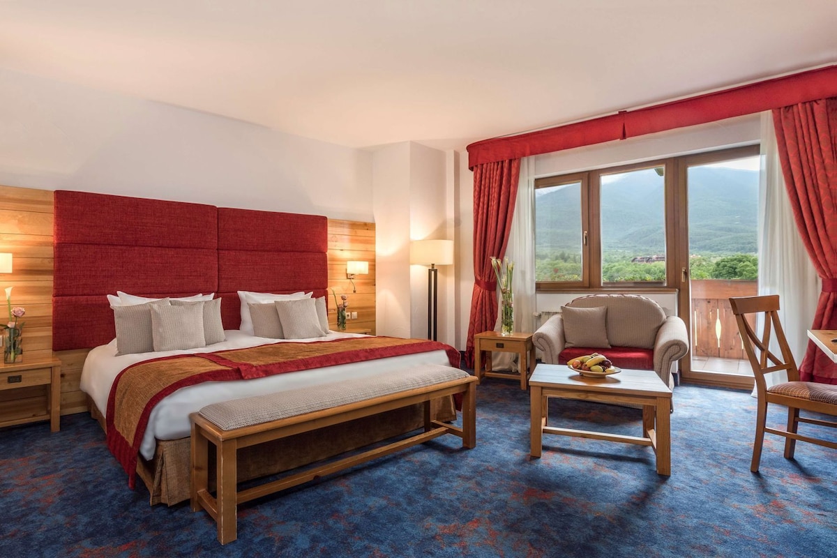 A hotel room with a large bed, a wooden bench, a small table with fruit, a sofa, and a view of the outdoors.