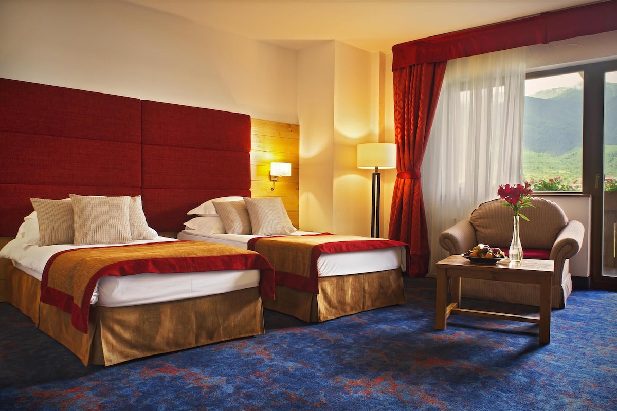 A hotel room with two beds, a sitting area, a lamp, and a view of mountains.