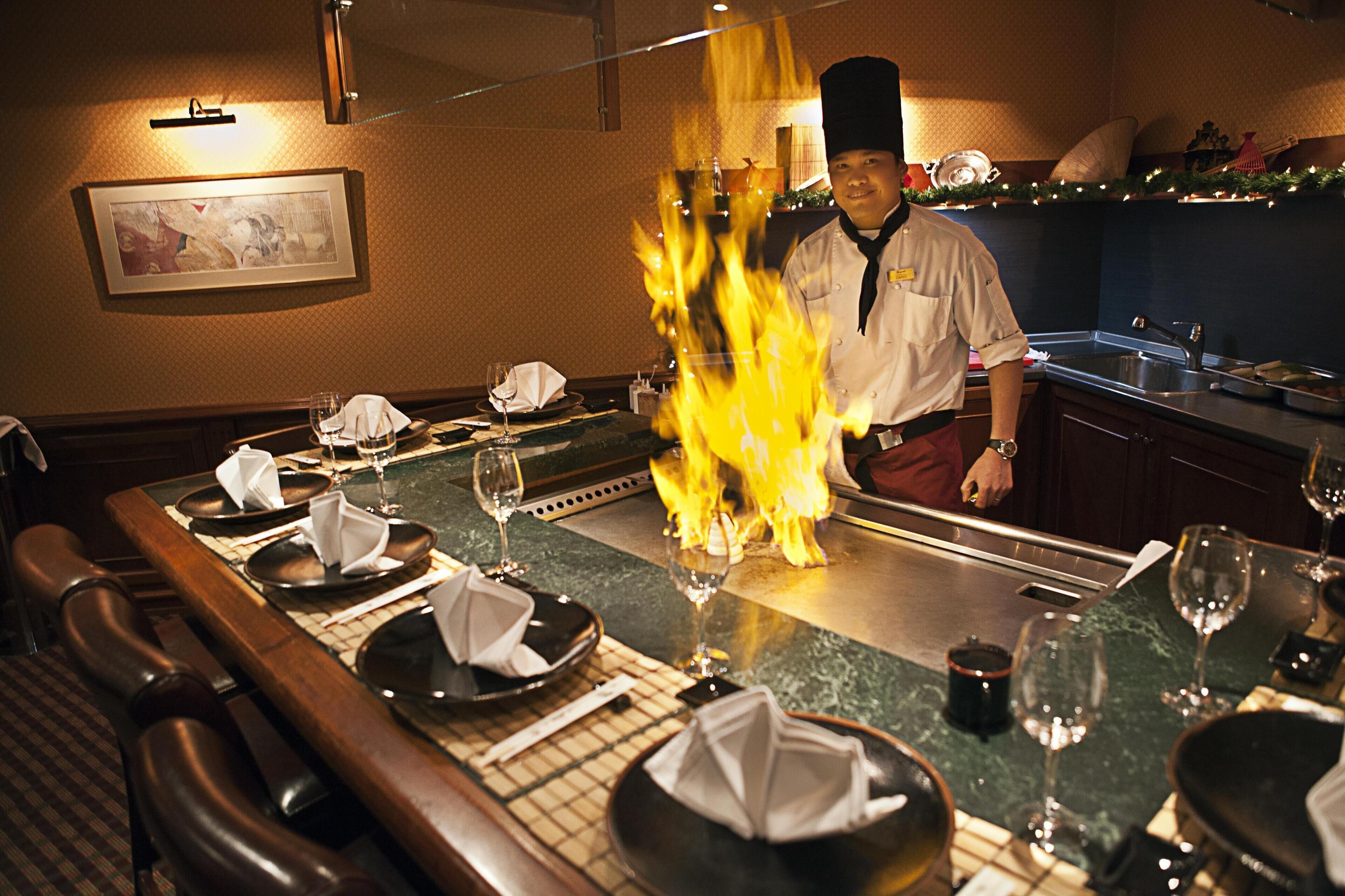 Un restaurant où une table est dressée pour le repas, un chef prépare un plat en le flammant, et un tableau encadré orne le mur.