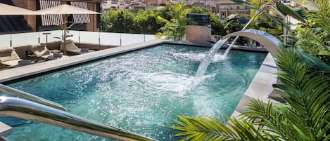 A rooftop pool with clear water, a waterfall feature, and a cityscape view in the background.
