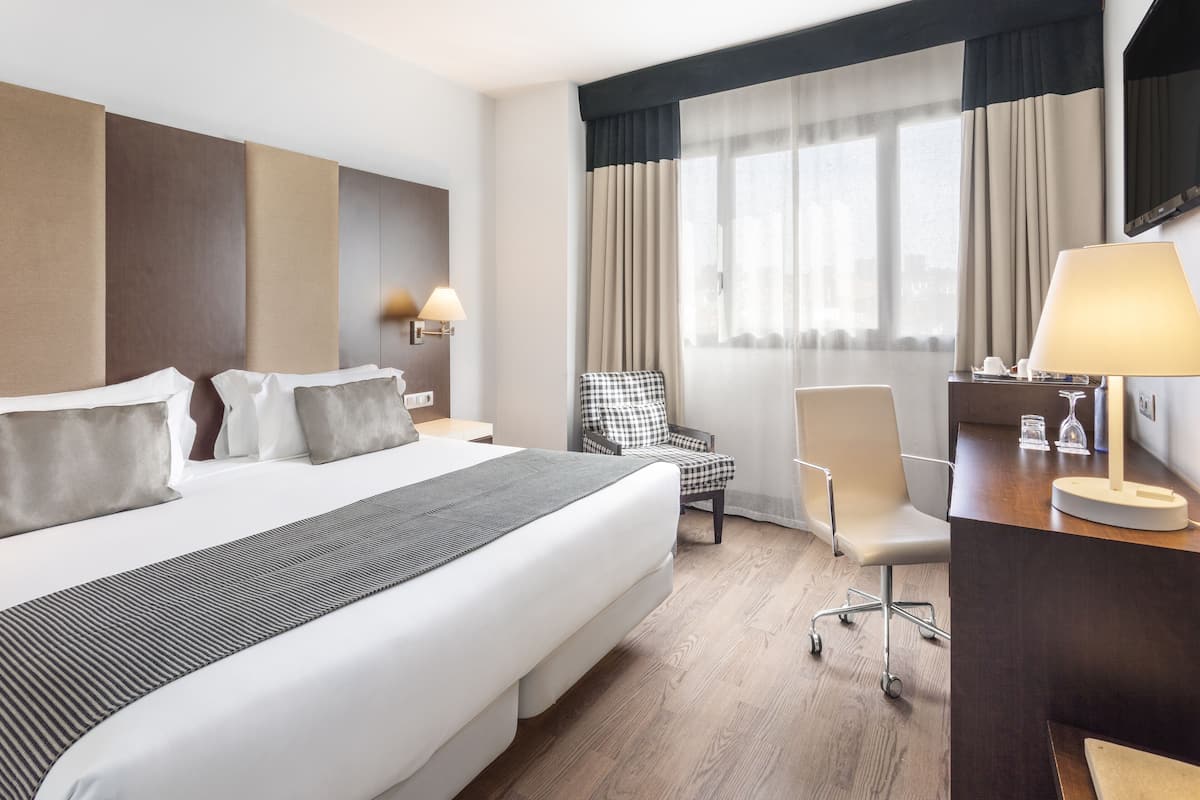 Una habitación de hotel con una cama grande, dos sillas, un escritorio con una lámpara y vistas a la ciudad a través de la ventana.