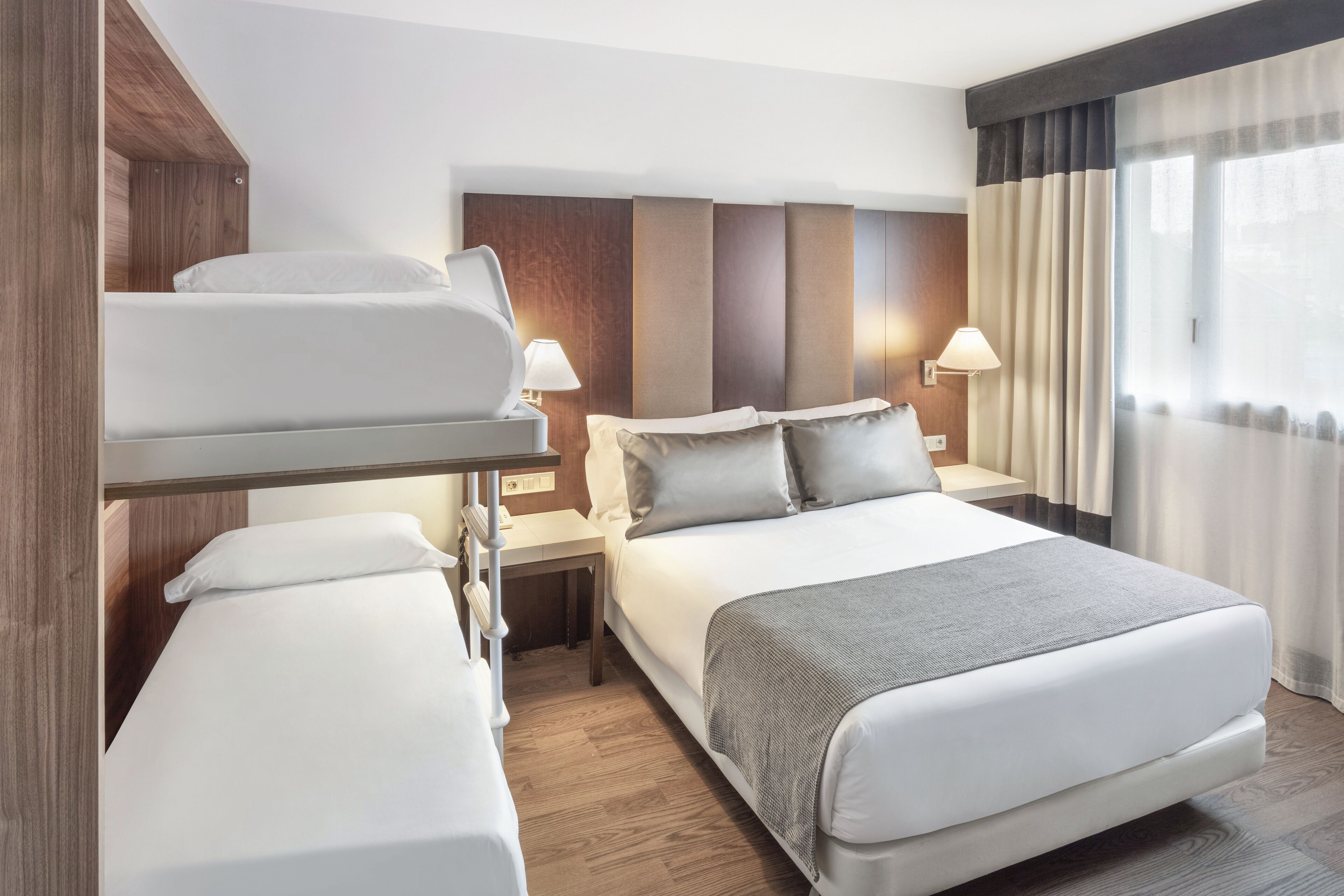 Habitación de hotel con literas, una cama individual, una mesita de noche con lámpara y vistas al exterior a través de una ventana con cortinas.