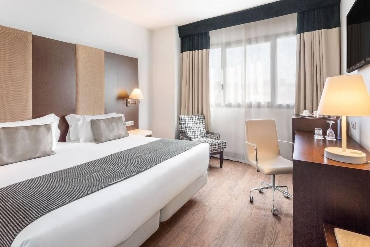 Une chambre d’hôtel avec un grand lit, deux chaises, un bureau et une fenêtre avec des rideaux.