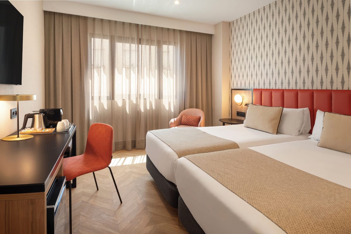 Habitación de hotel con dos camas, un escritorio con silla, televisor y ventana con cortinas.