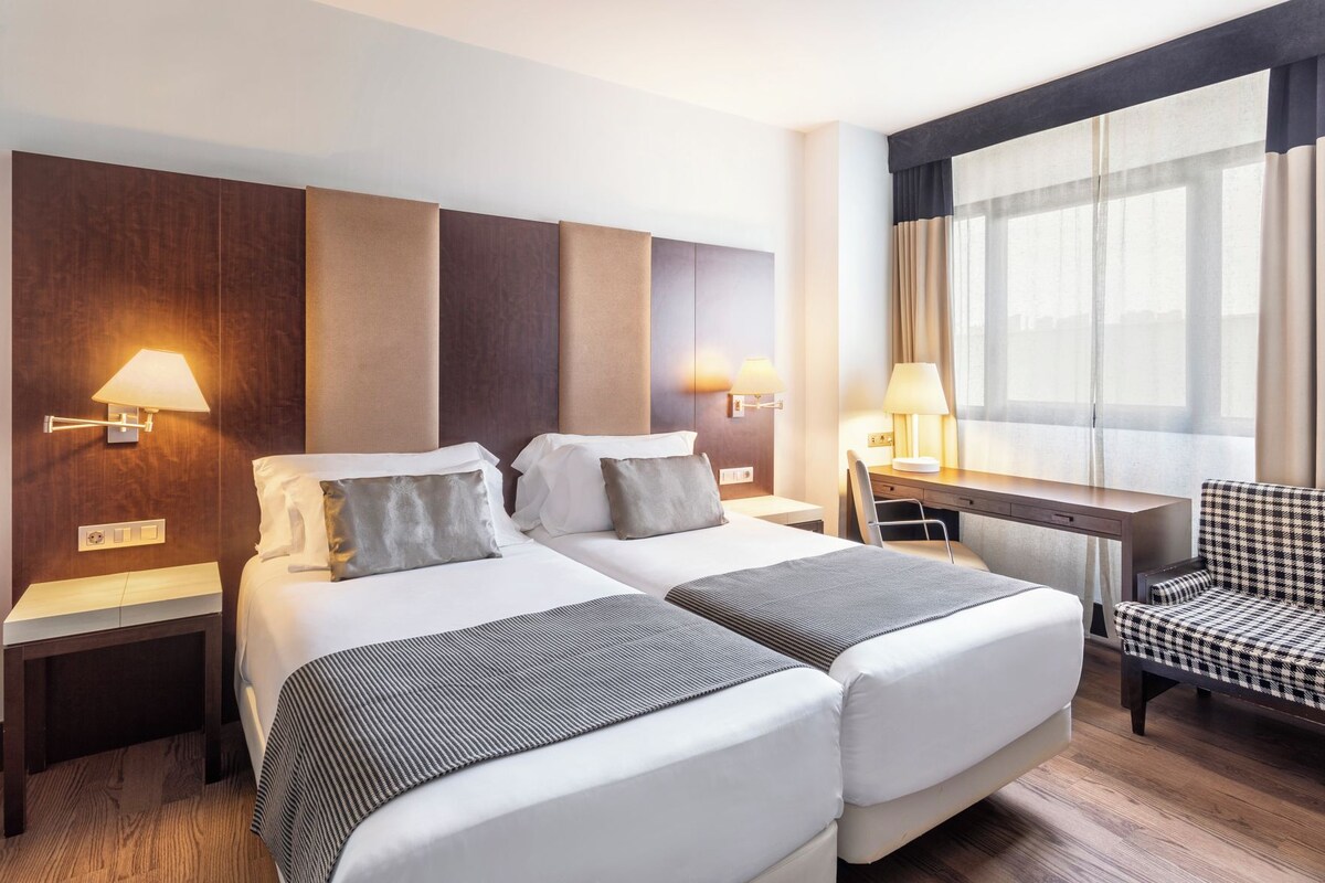 Une chambre d’hôtel avec deux lits, un bureau, une chaise et une grande fenêtre avec des rideaux.