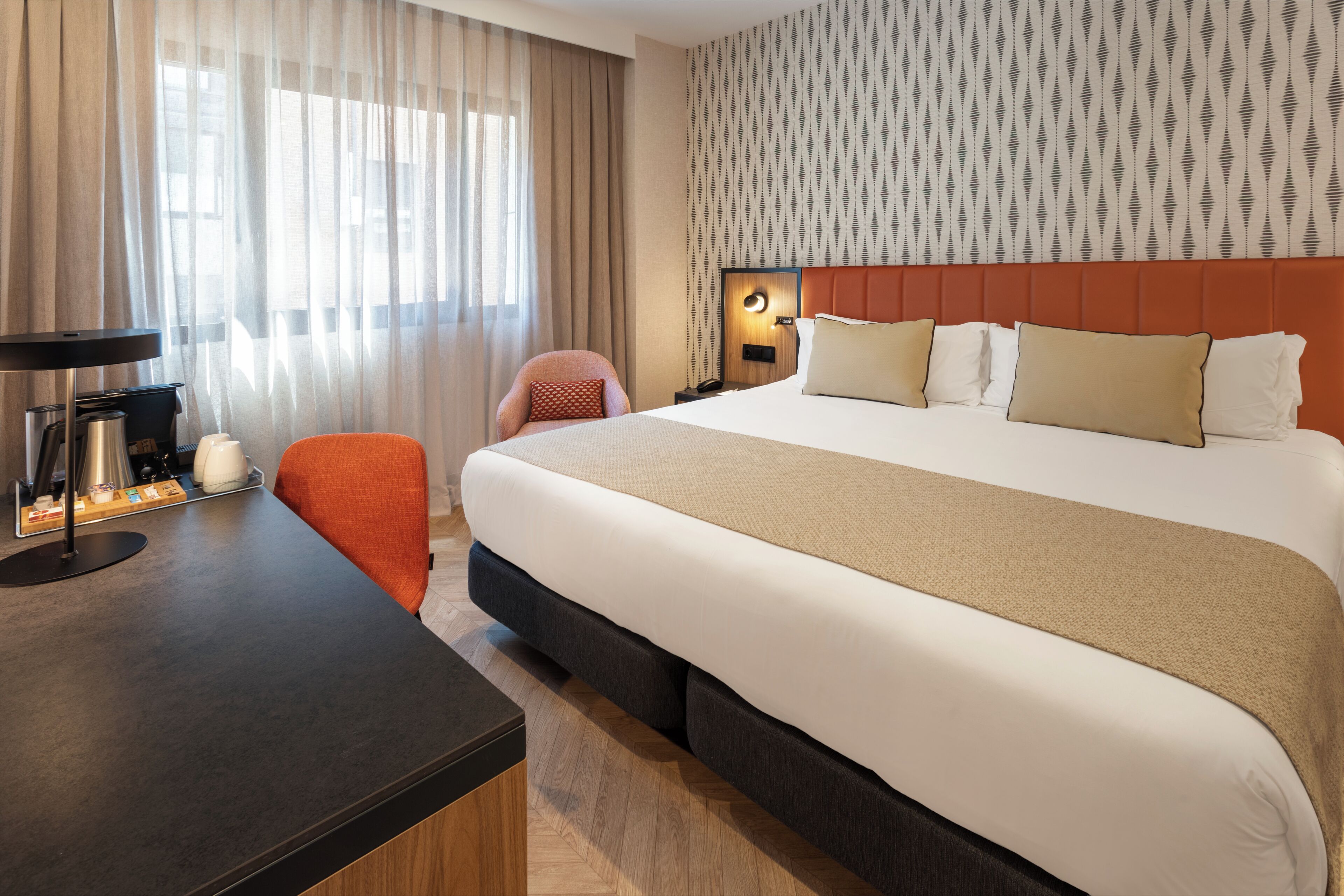 A hotel room with a large bed, a desk with a lamp, a chair, and a patterned wall.