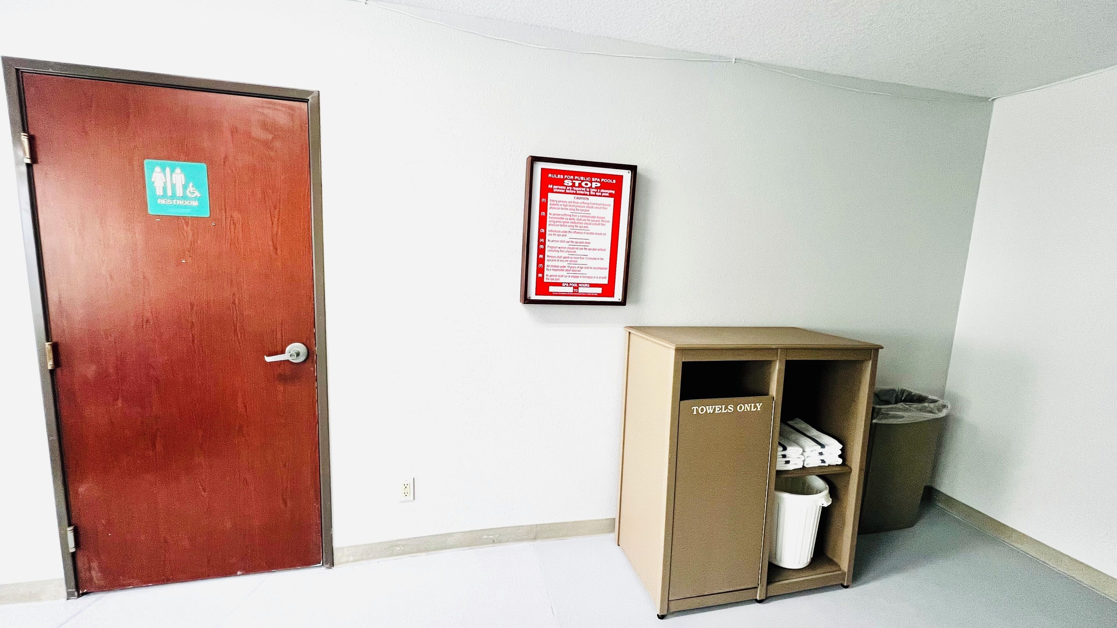 A hotel room with a wooden door, a sign indicating a restroom, a framed rule sign for public pools, and a storage unit labeled 'TOWELS ONLY'.