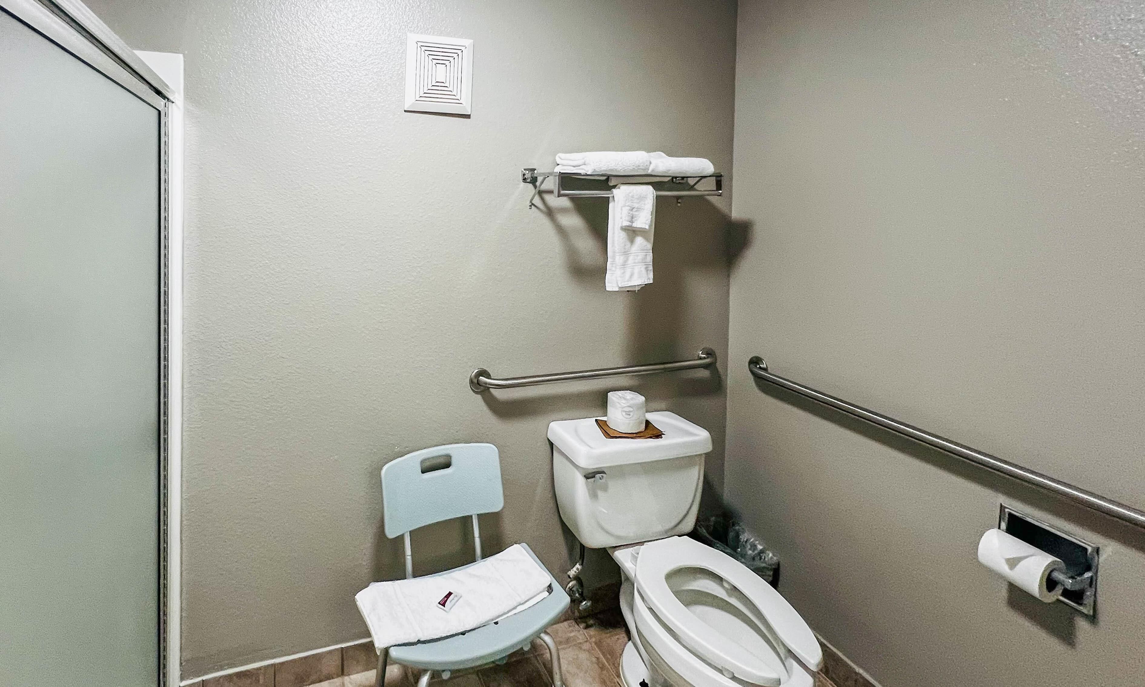 A small, single-occupancy bathroom with a toilet, a chair, a towel rack, and a handrail.