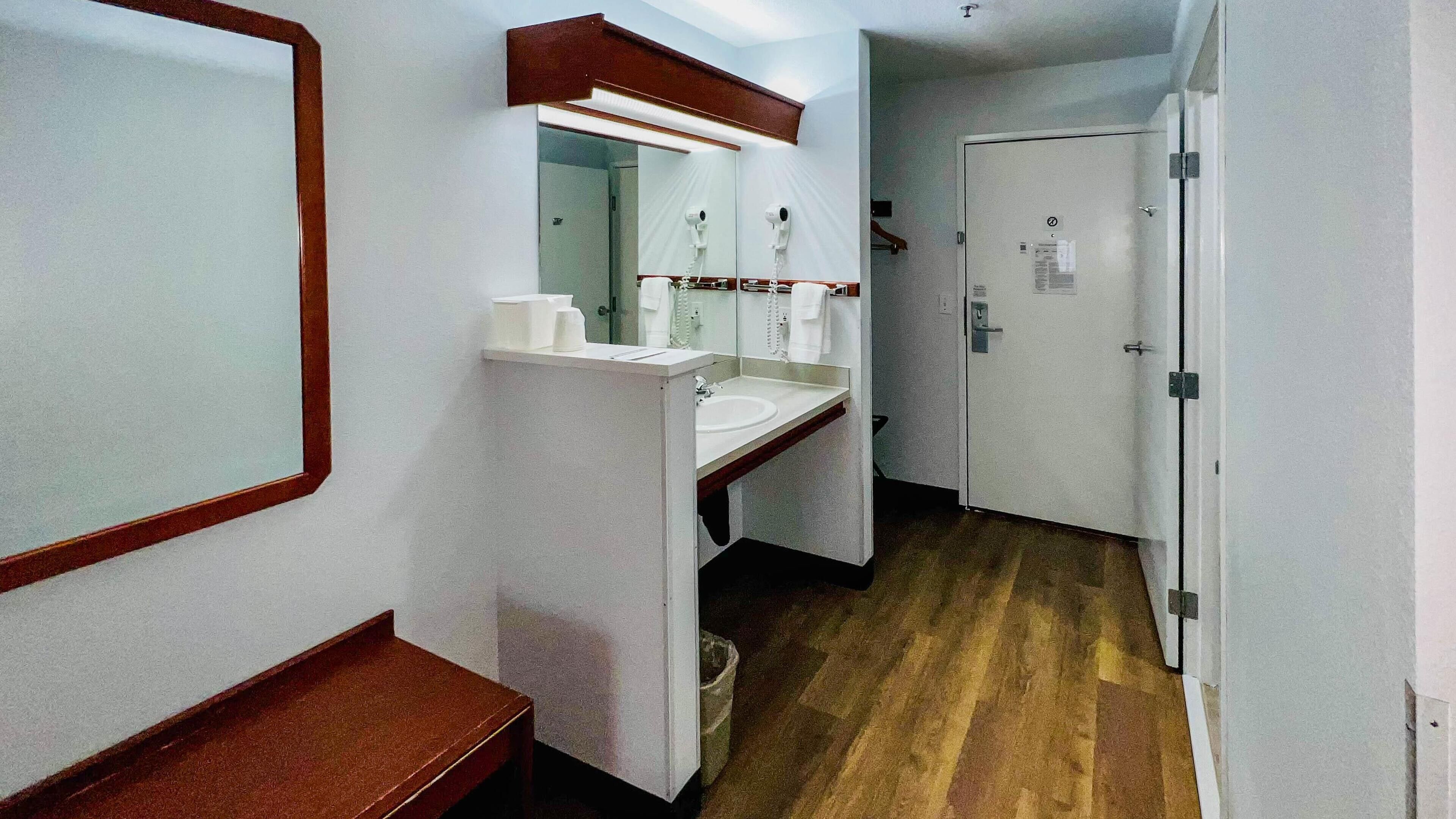 A narrow hallway with a white sink, mirror, and towel rack. A wooden bench is on the left, and a door is on the right.