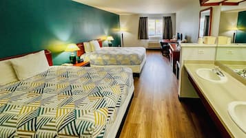 A hotel room with two beds, a desk, a computer, a desk lamp, a mirror, and a bathroom with a sink and mirror.