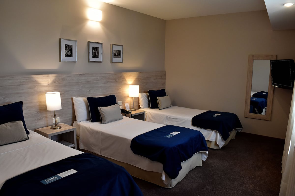 Une chambre d’hôtel avec trois lits, chacun recouvert d’une couverture bleue, avec du linge de lit blanc et une table de chevet avec une lampe. Des tableaux encadrés ornent le mur, et un miroir est accroché sur le côté droit.