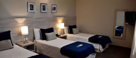 A hotel room with three beds, each with a blue cover, white bedding, and a nightstand with a lamp. There are framed pictures on the wall and a mirror on the right side.