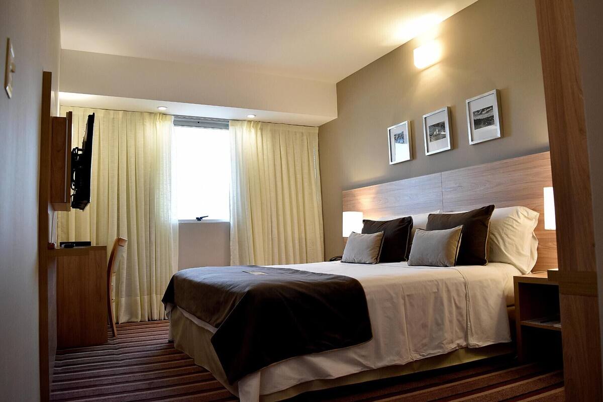 Une chambre d’hôtel avec un lit, une table de chevet, une fenêtre avec des rideaux et un téléviseur fixé au mur.