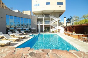 A swimming pool with lounge chairs, a bar area, and a building with a staircase and large windows.