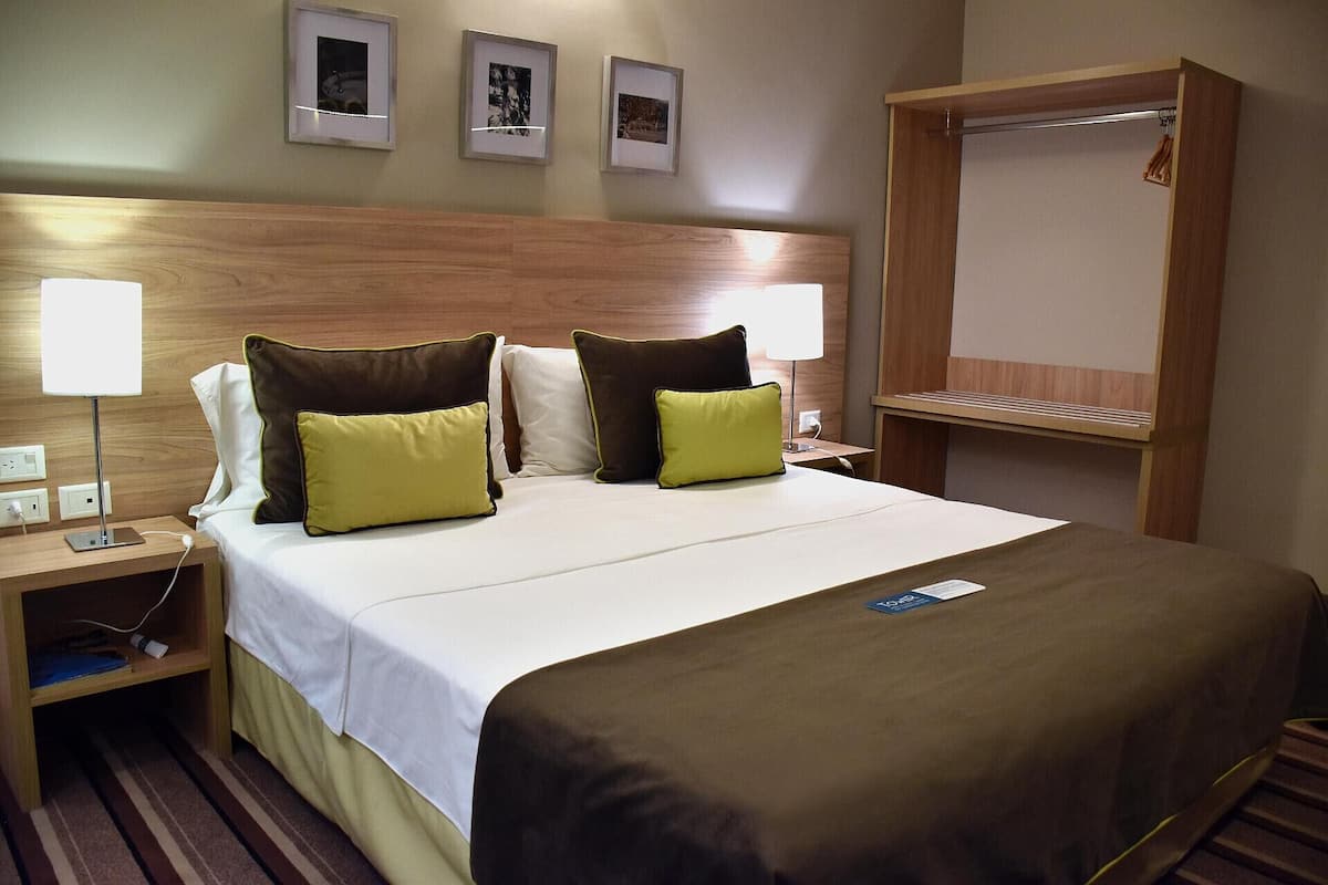 Une chambre d’hôtel avec un grand lit, deux lampes de chevet, une tête de lit en bois, une table de chevet et une armoire.