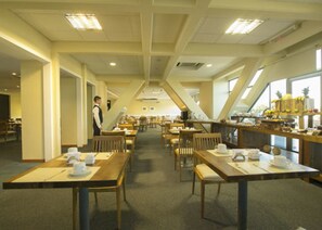 A spacious dining area with multiple tables and chairs, a waiter standing by the counter, and a view of the outdoors through large windows.