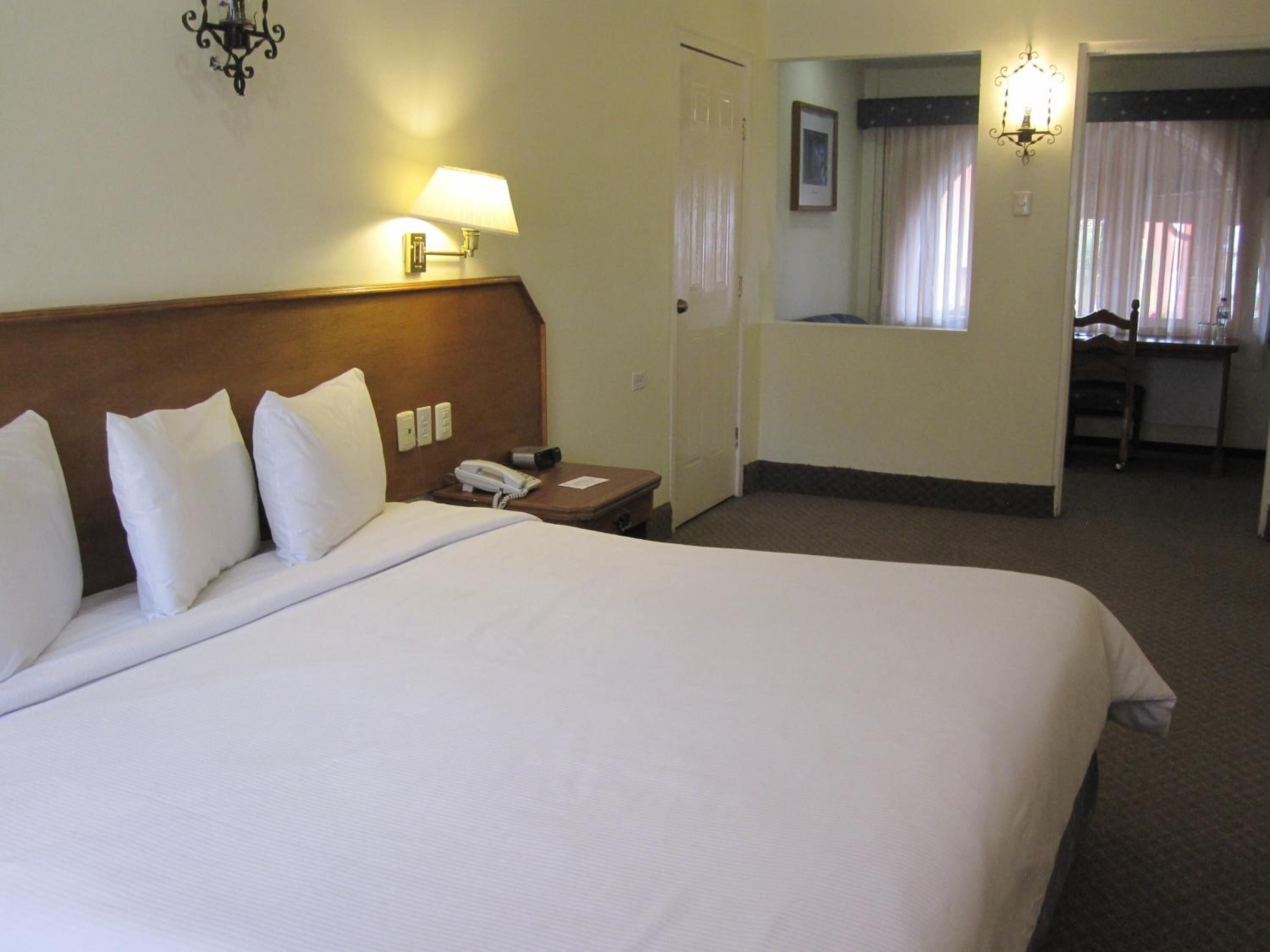 A hotel room with a large bed, a bedside table with a lamp, a phone, and a door leading to a bathroom.