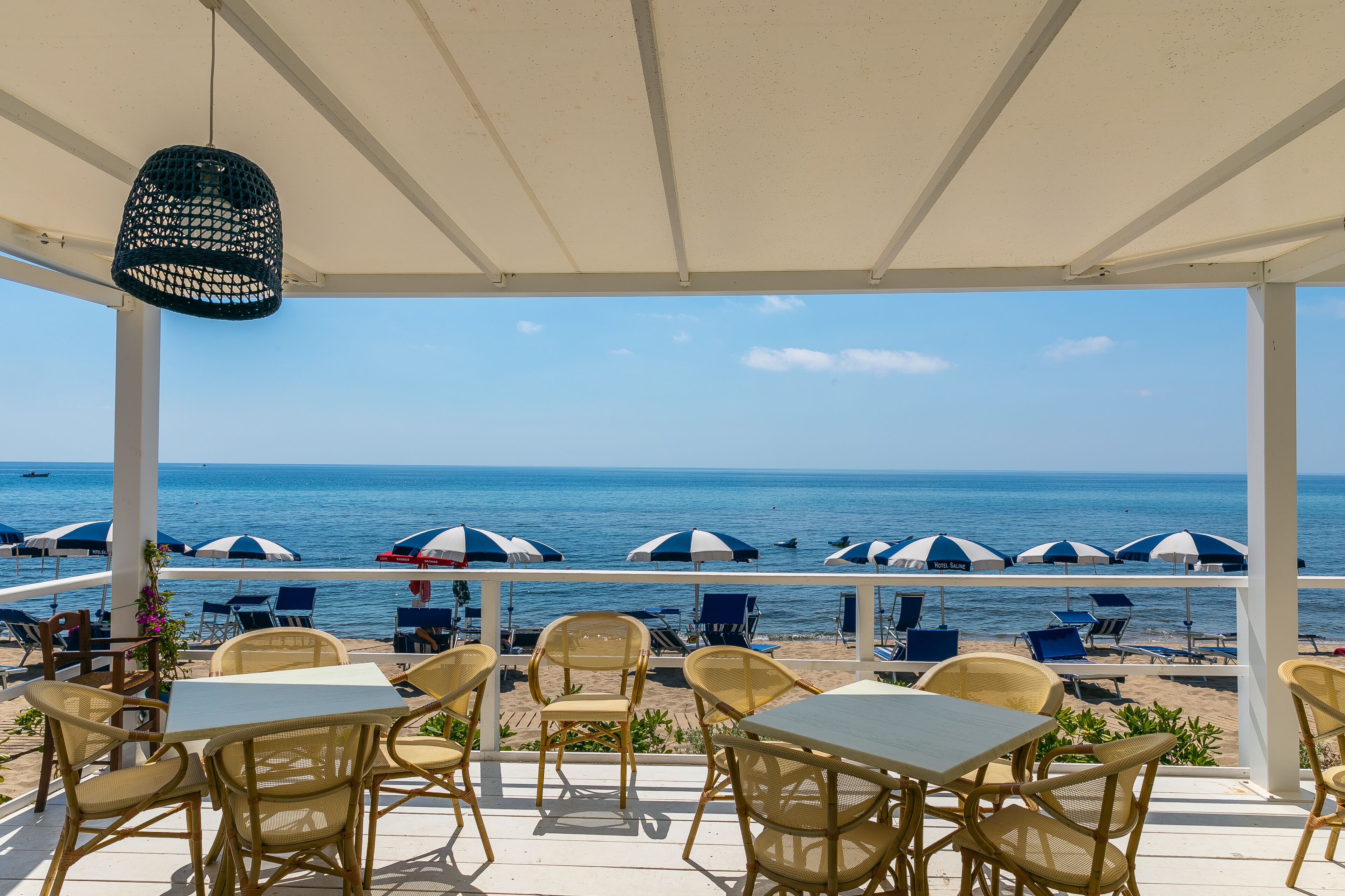 Un'area salotto esterna riparata con sedie e tavoli in vimini, affacciata su una spiaggia con ombrelloni e il mare.