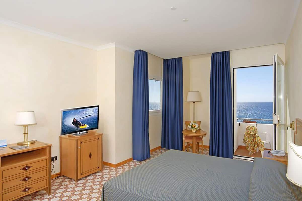 Une chambre d’hôtel avec un lit, un bureau, une télévision, une fenêtre avec des rideaux et une vue sur la mer.