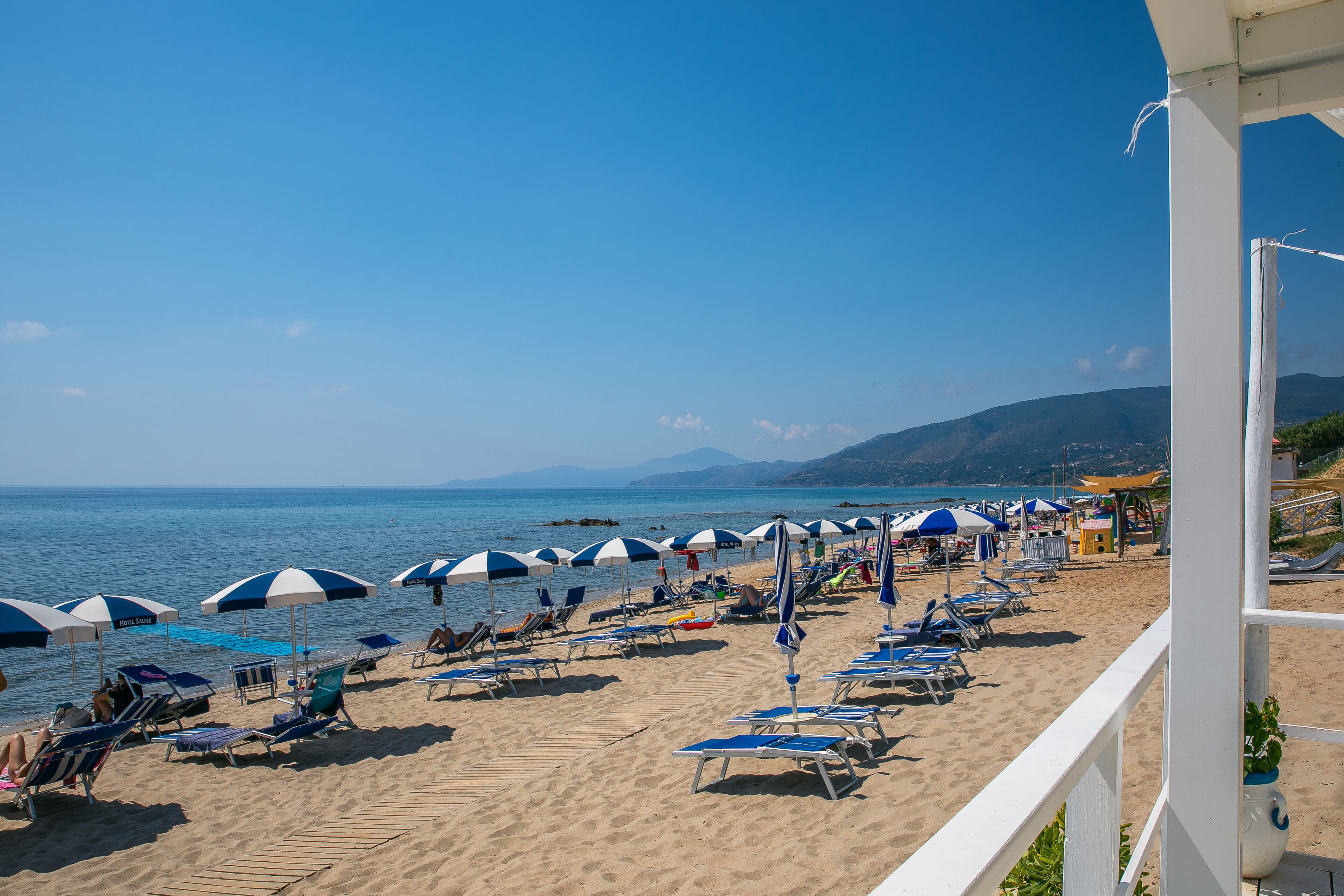 A beach with rows of sun loungers and umbrellas, a clear sky, and a view of the sea and mountains in the background.