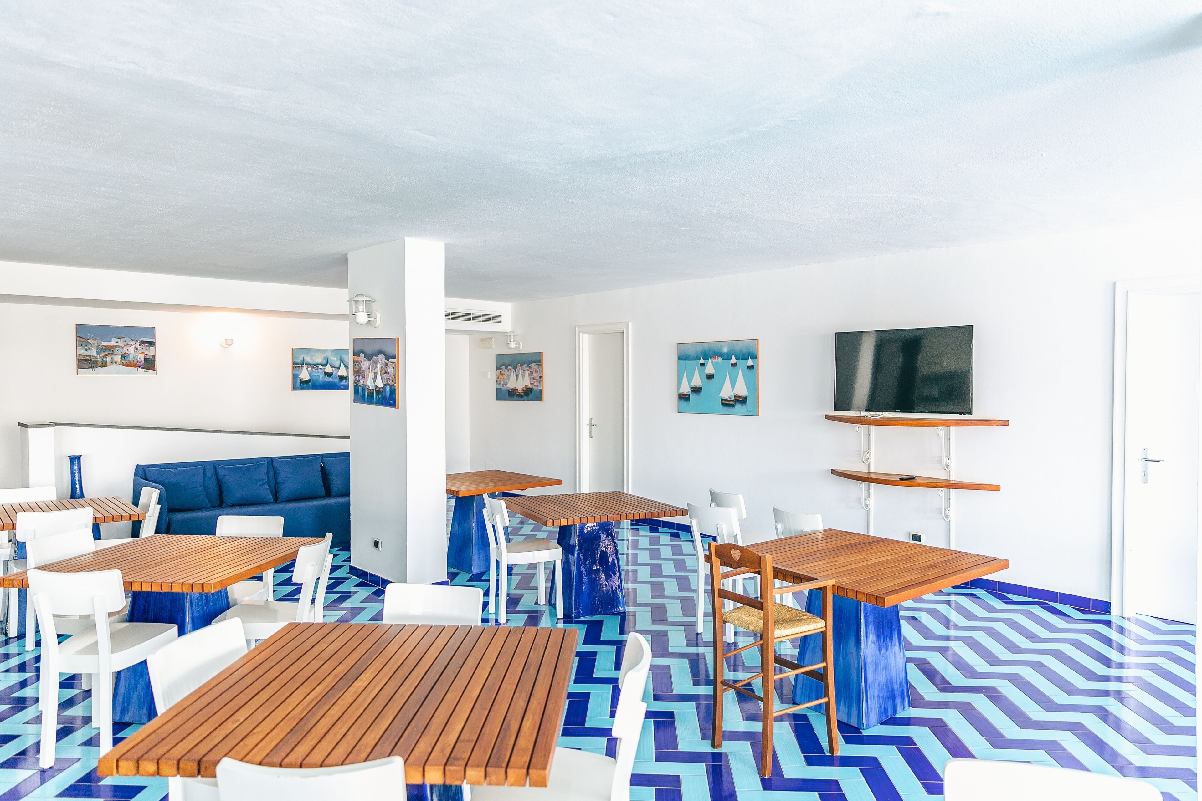 Un espace repas moderne avec des tables en bois et des chaises blanches, un sol à motifs bleus et des œuvres d’art murales sur le thème de la marine.