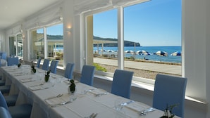 Ein langer Esstisch mit Stühlen, gedeckt für eine Mahlzeit, mit Blick auf einen Strand mit Sonnenschirmen und das Meer.