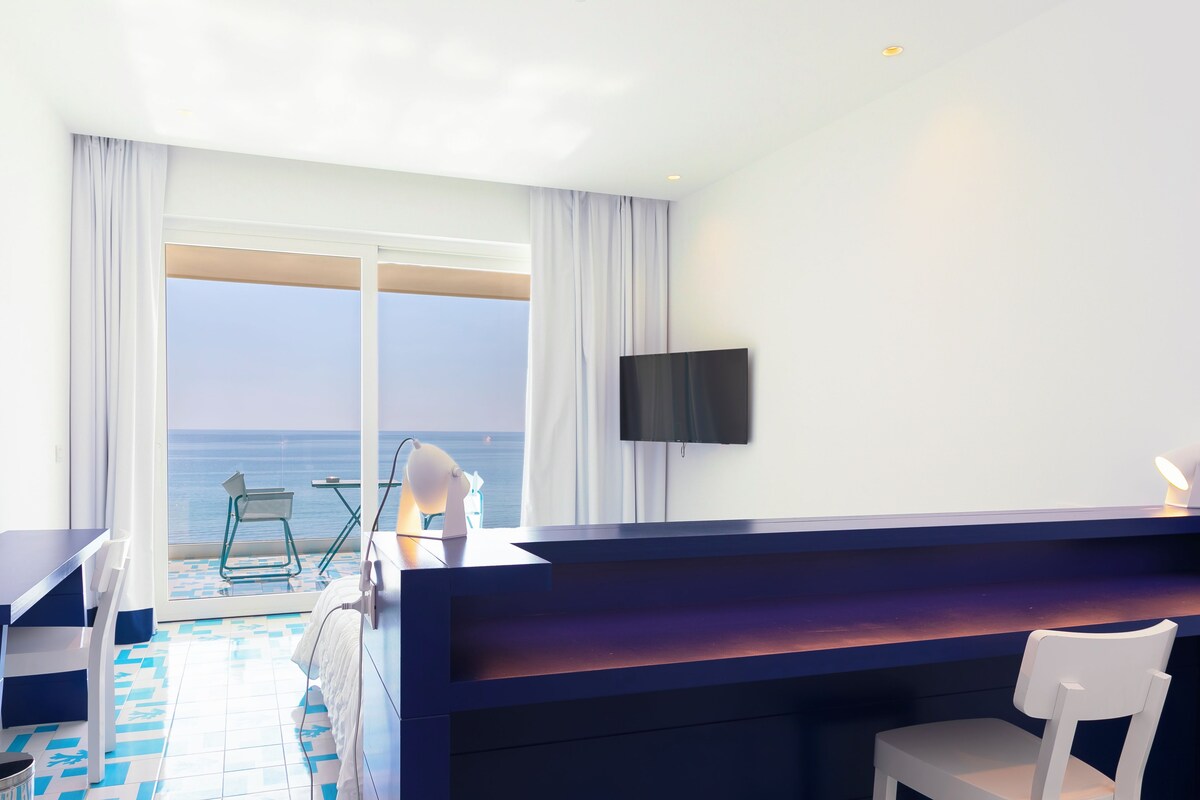 Ein Hotelzimmer mit Balkon, Schreibtisch, Stuhl, Fernseher und Meerblick.