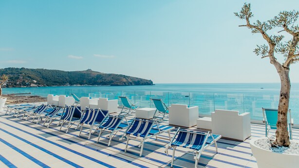 Ein Poolbereich mit blau-weißen Liegestühlen und weißen Sofas, mit Blick auf das klare, blaue Meer und einen entfernten, grünen Hügel.